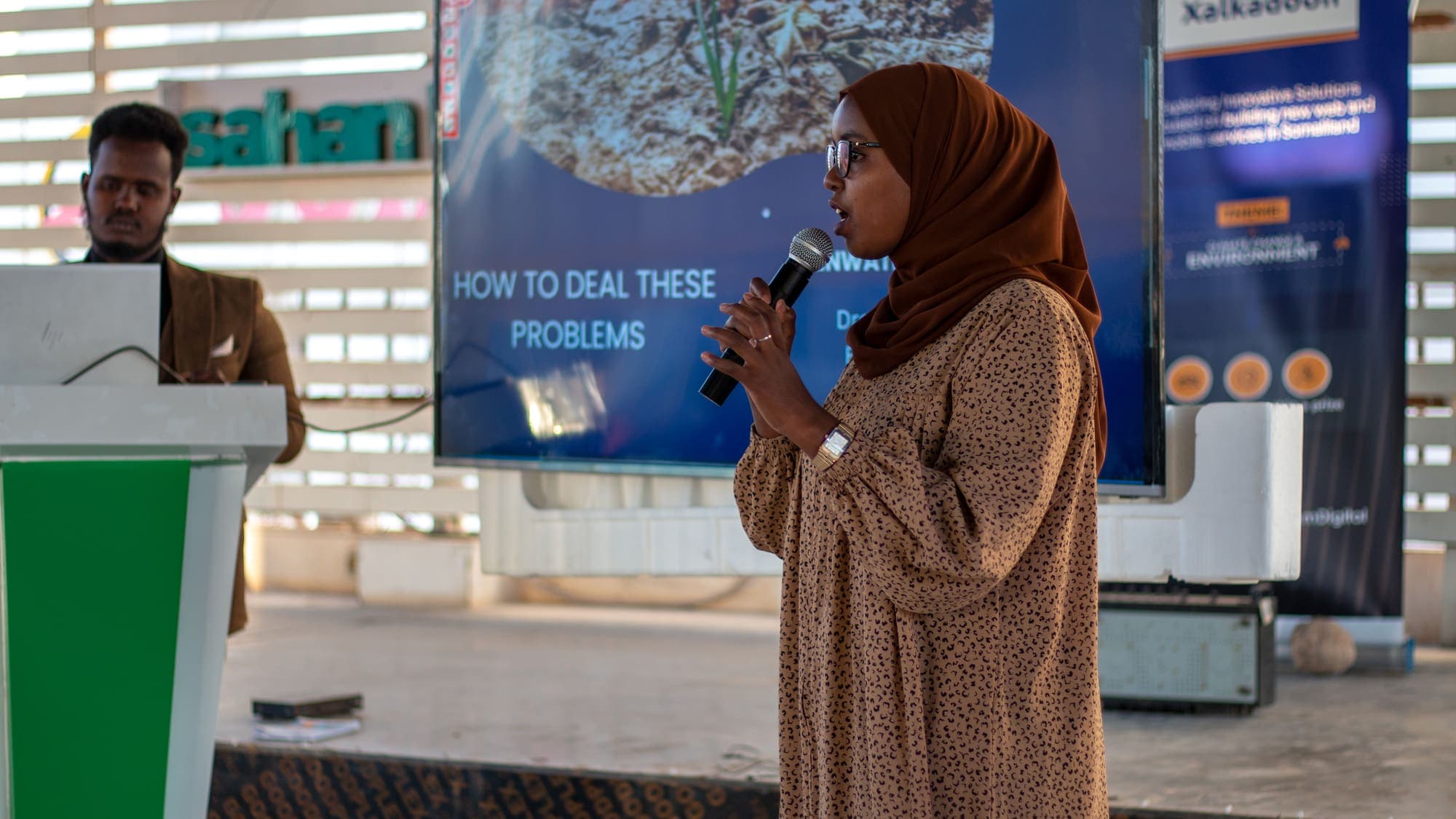 A presenter with a microphone stands beside her slides