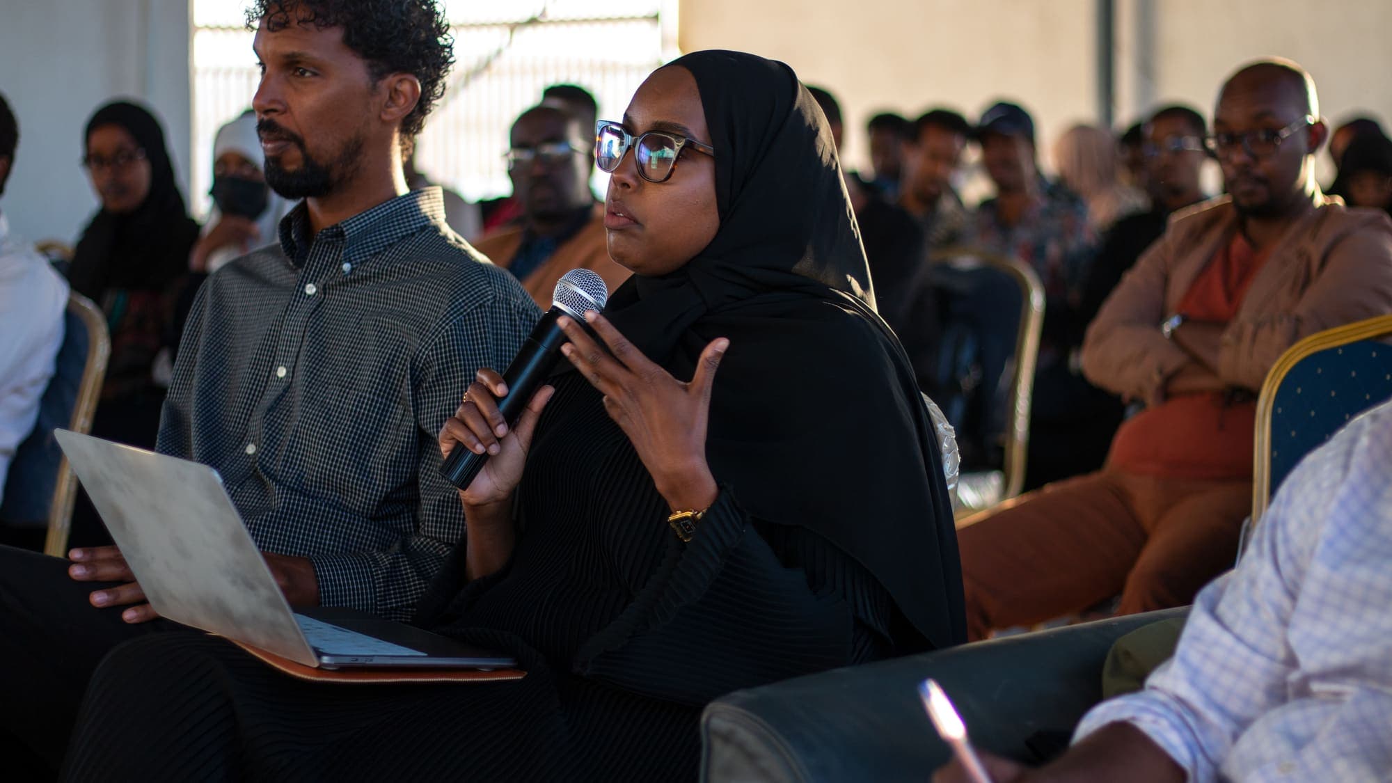 A woman with a laptop on her knees speaks into a microphone from her seat