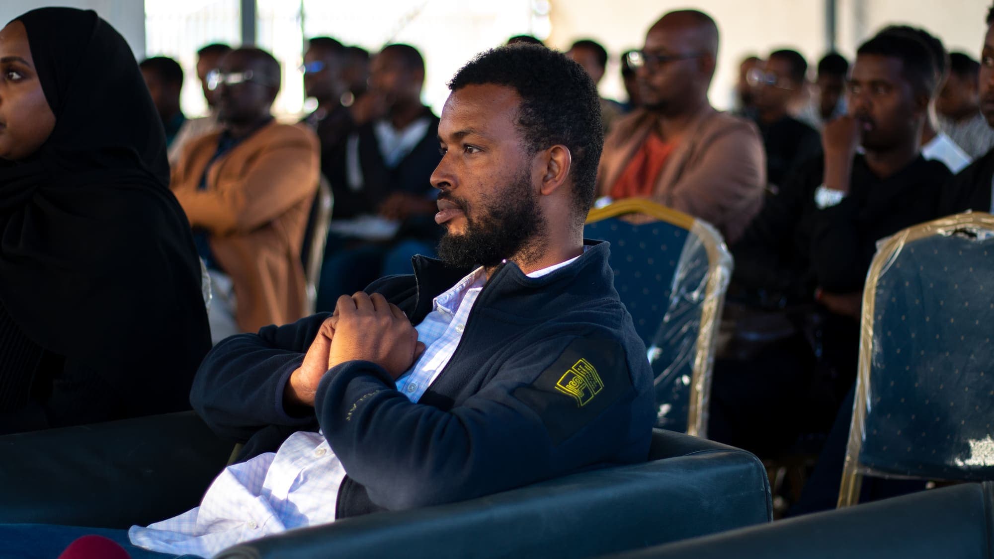 A man seated on a sofa at the front listens with his hands clasped